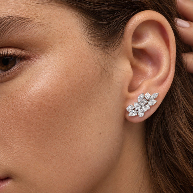 A close-up of a woman's ear wearing a cluster stud earring made of clear gemstones in a silver-toned setting. The earring is shaped like a small floral or leafy cluster and is photographed against skin tones with brown hair.