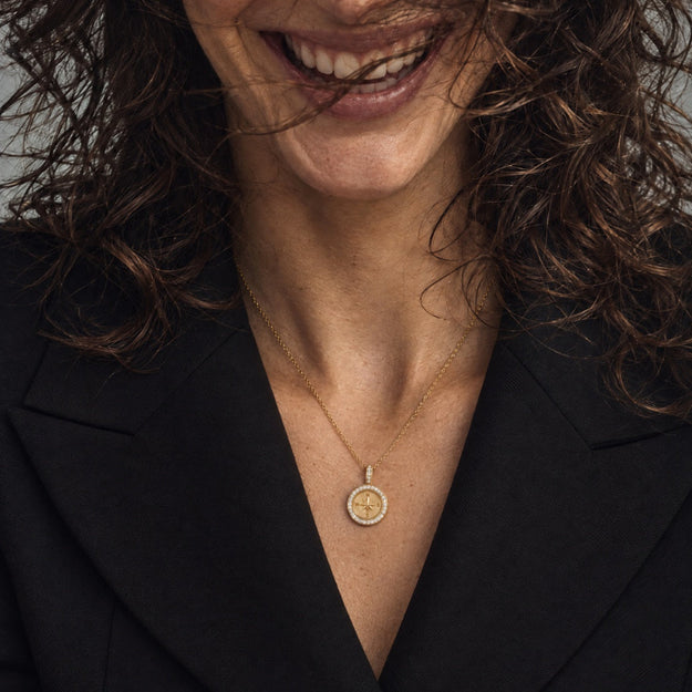 A close-up lifestyle shot of a woman wearing a delicate gold pendant necklace with a circular sun design, styled with a black blazer.