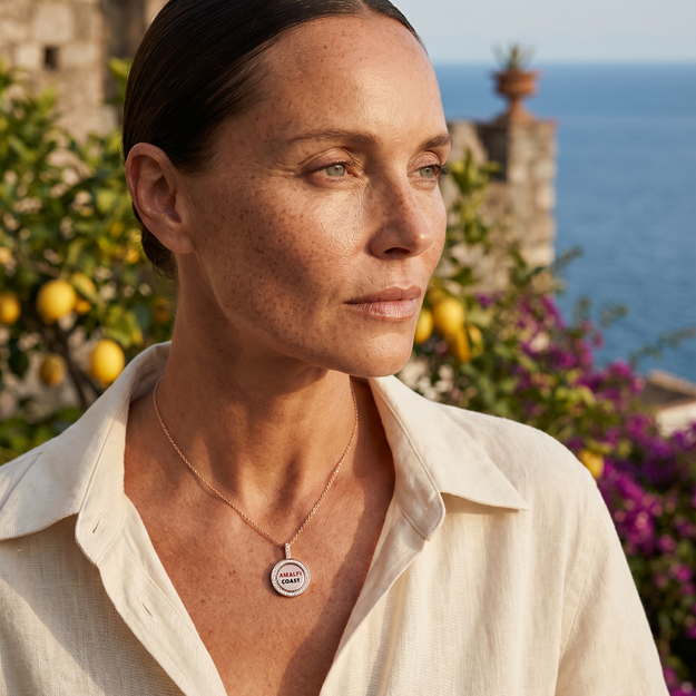 Woman wearing a necklace with a circular pendant, standing in front of a scenic background with ocean and castle.