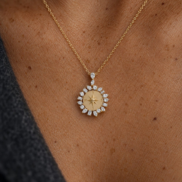A close-up of a gold necklace with a compass rose pendant, worn on a person's neck. The pendant features a gold center with a star design and is surrounded by small clear gemstones in a circular setting.