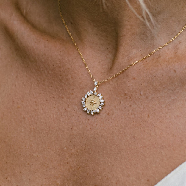 A close-up of a gold necklace with a circular compass pendant adorned with small clear stones, worn on a person's neck.