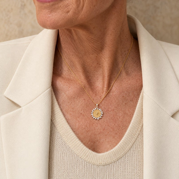 A close-up of a woman wearing a delicate gold necklace with a sunburst pendant, styled with a cream-colored knit top and blazer.