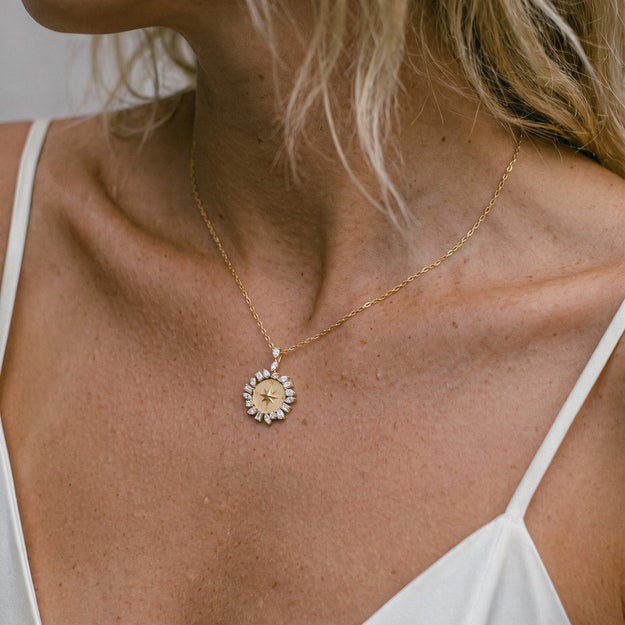 A close-up of a woman's neck and chest wearing a delicate gold chain necklace with a circular pendant featuring a gold center and a halo of small clear stones.