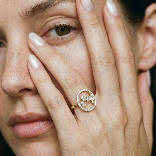 Close-up of a woman's face with one hand covering her eye, wearing a gold ring with pearls.