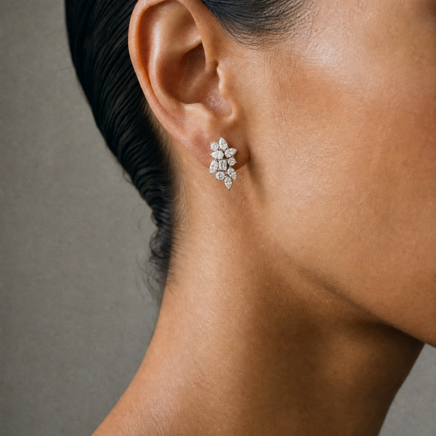 A close-up side profile of a model's ear wearing a small floral cluster stud earring with clear stones in a silver-toned setting, against a neutral gray background.